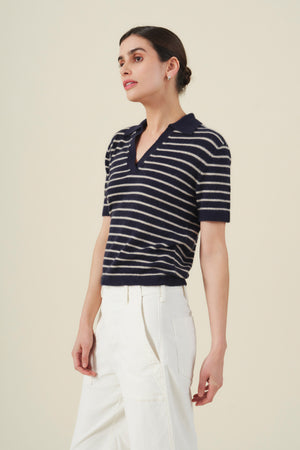 A person wearing the Velvet by Graham & Spencer YAEL Cashmere Sweater Polo—a navy blue and white striped short-sleeve knit top with subtle striped detailing—paired with white pants stands against a plain light background.