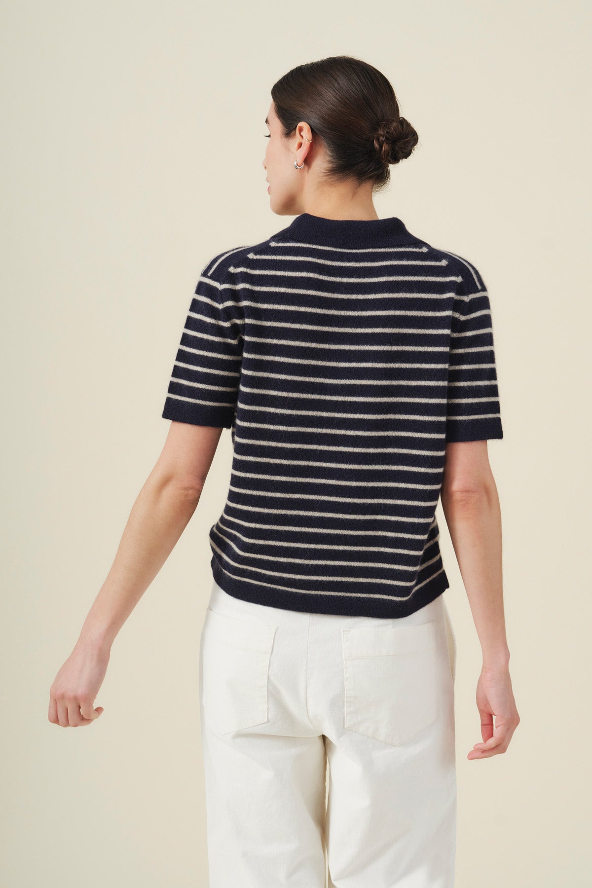A person with dark hair in a low bun is shown from the back against a plain background, wearing the Velvet by Graham & Spencer YAEL Cashmere Sweater Polo—a navy short-sleeve top with striped detailing—paired with white pants.-41976495407297