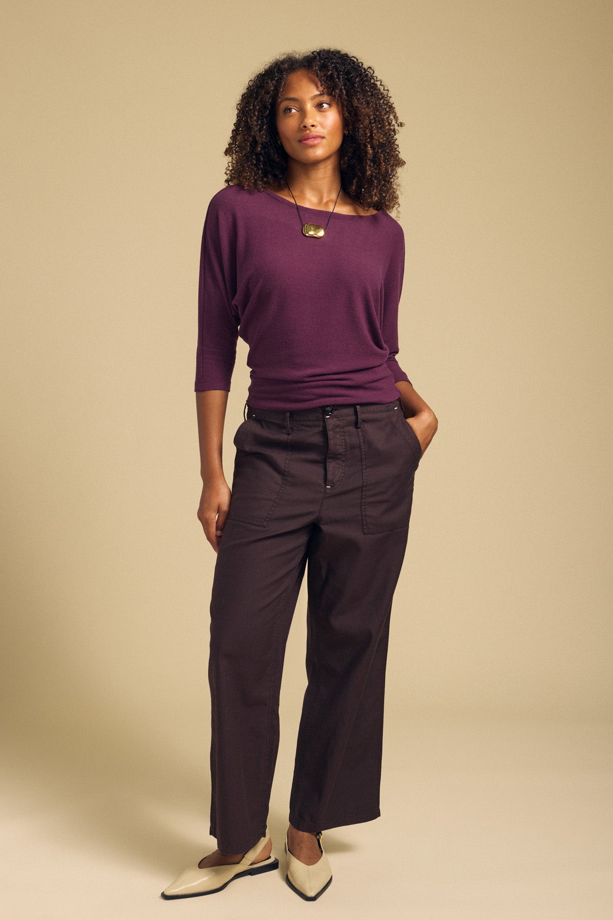 A woman stands against a beige background wearing a purple top, the JACLYN PANT by Velvet by Graham & Spencer in wide leg cotton sateen, cream pointed shoes, and a gold necklace.