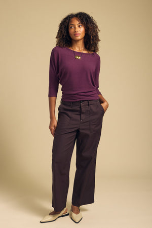 A woman stands against a beige background wearing a purple top, the JACLYN PANT by Velvet by Graham & Spencer in wide leg cotton sateen, cream pointed shoes, and a gold necklace.