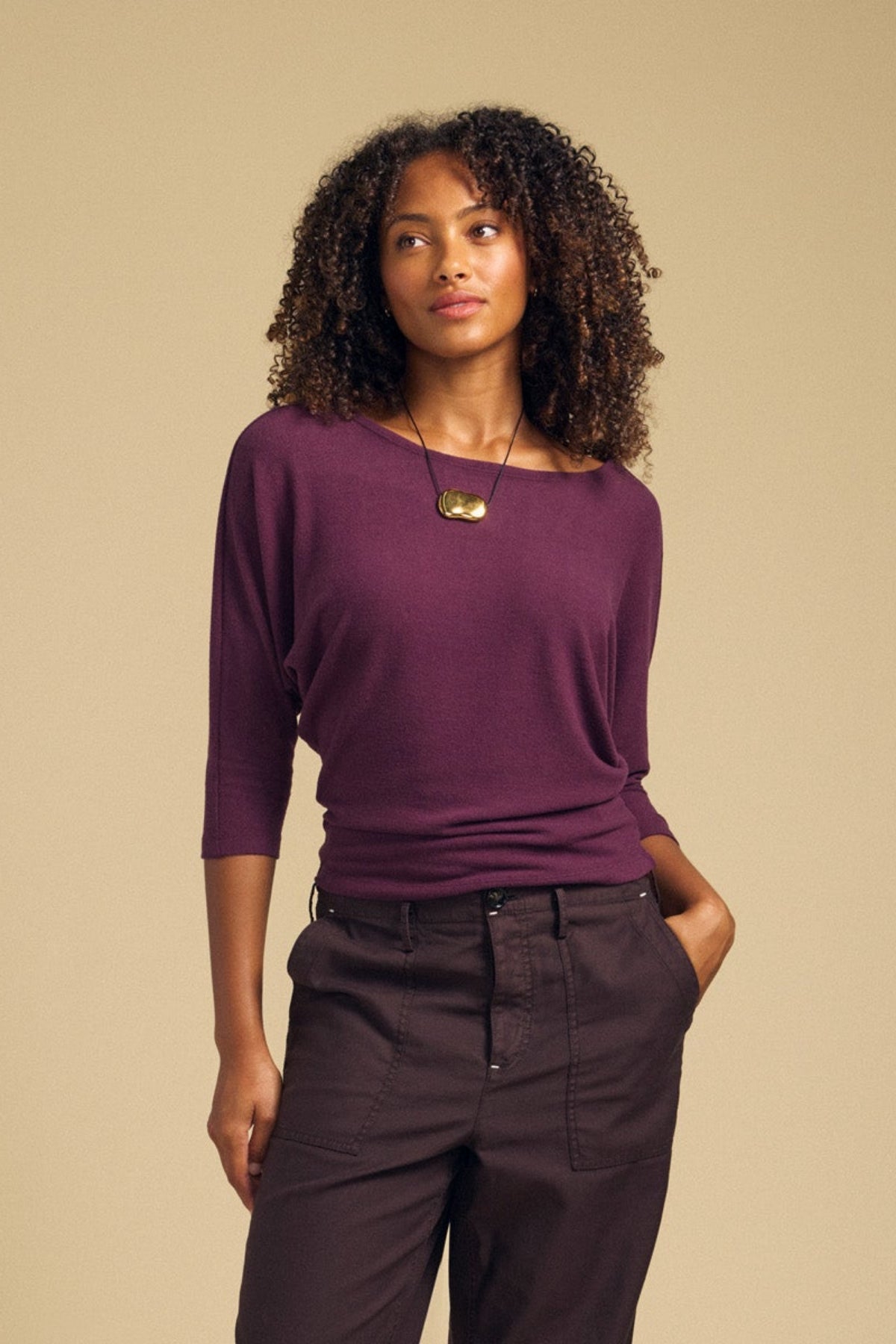 A woman with curly hair wears the CECILIA TOP by Velvet by Graham & Spencer—a relaxed fit, purple brushed knit top—paired with dark pants, standing with one hand in her pocket against a plain beige background.-42063734440129