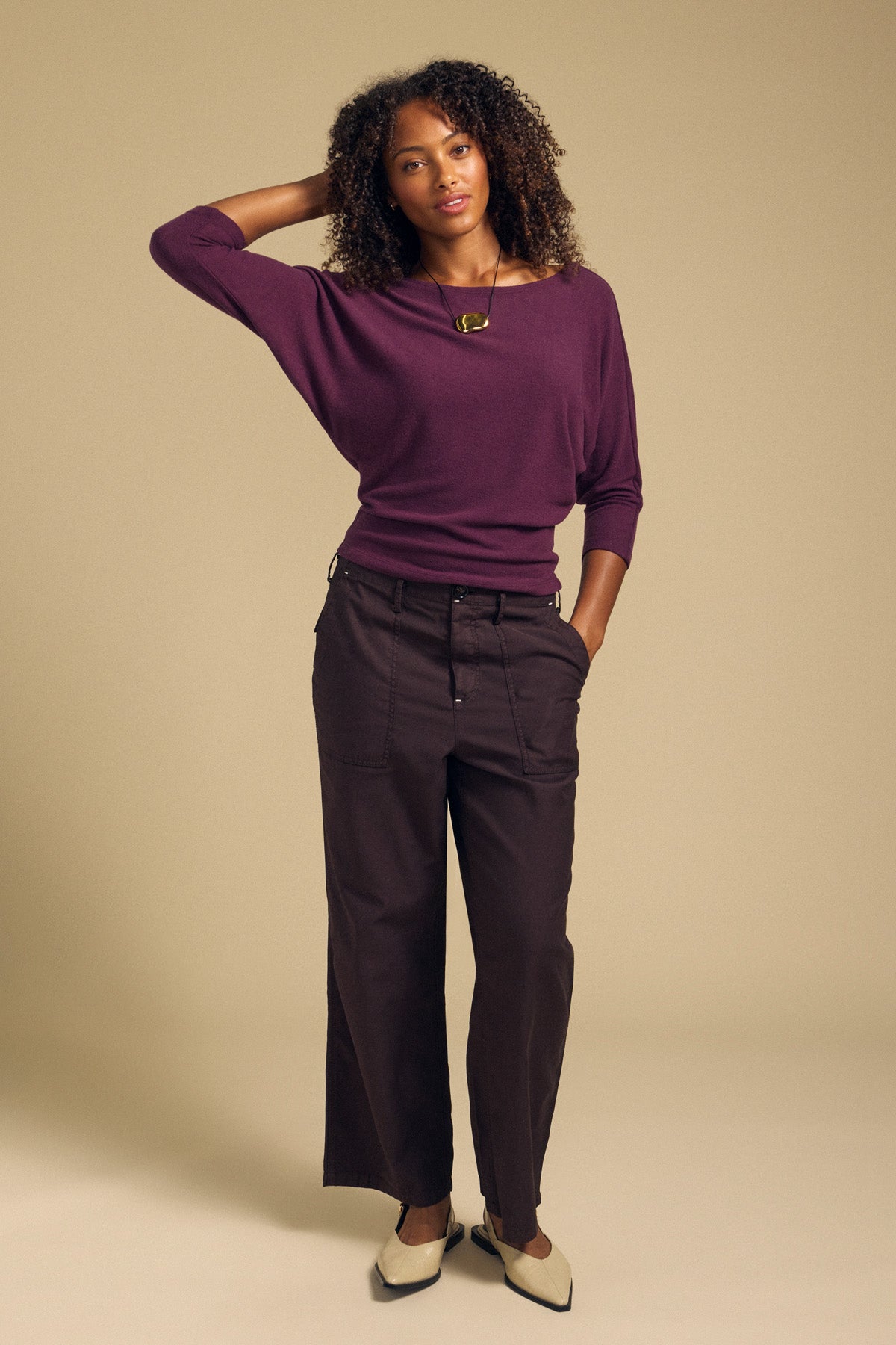 A woman stands against a beige background, wearing a long-sleeve purple cropped top, the JACLYN PANT by Velvet by Graham & Spencer—dark wide-leg pants crafted from brushed cotton sateen—paired with light shoes and a necklace, with one hand touching her hair.-41980268609729