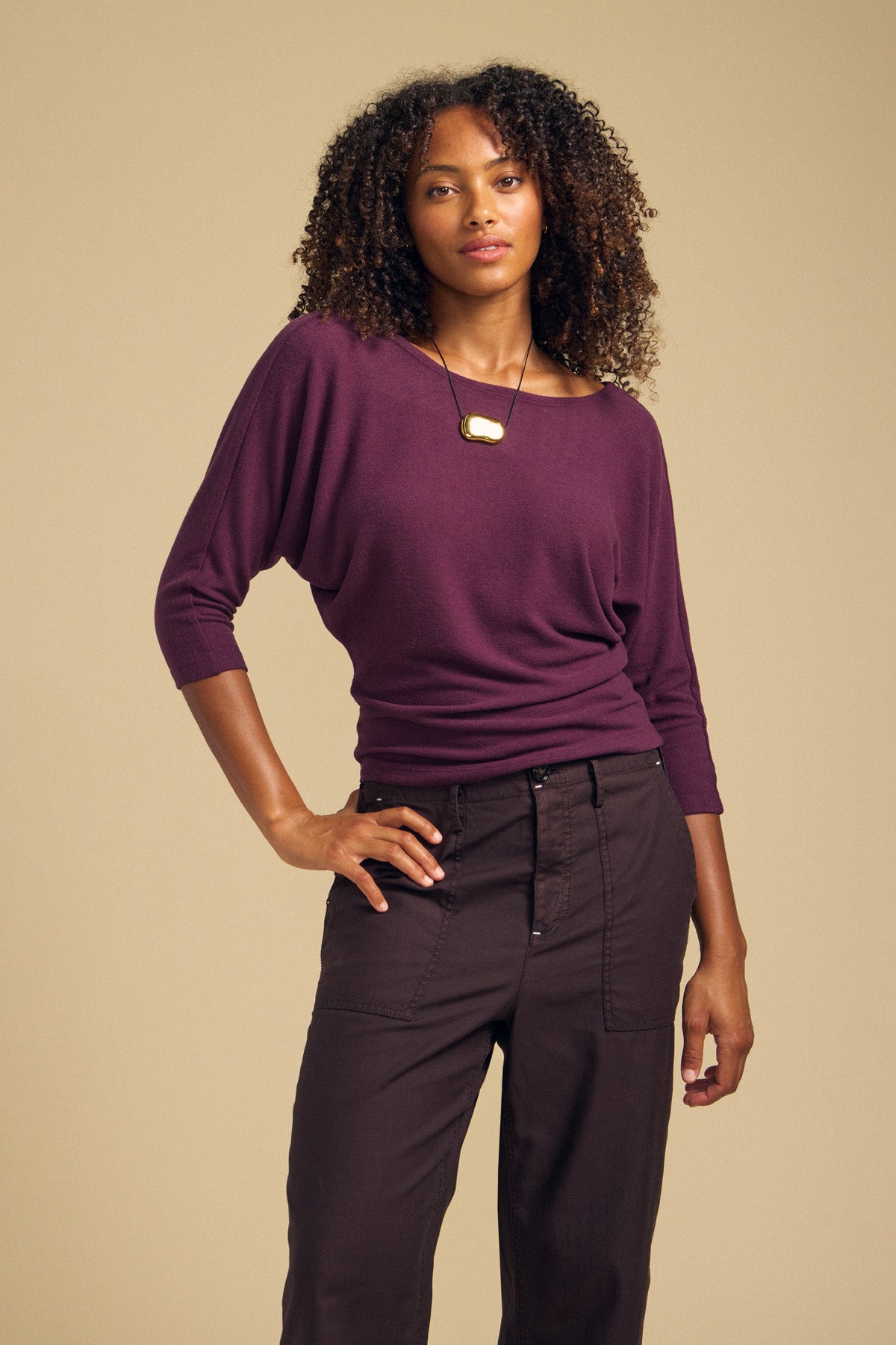 Standing against a beige background, the person is wearing the Velvet by Graham & Spencer CECILIA TOP—a purple brushed knit long-sleeve top with dolman sleeves—paired with dark pants and a pendant necklace, with one hand resting on their hip.