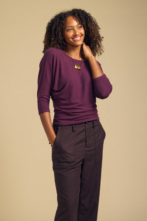 A woman stands against a beige background wearing the CECILIA TOP by Velvet by Graham & Spencer—a purple brushed knit long-sleeve with subtle dolman sleeves—paired with dark pants and a gold pendant necklace, one hand in her pocket and the other touching her hair.