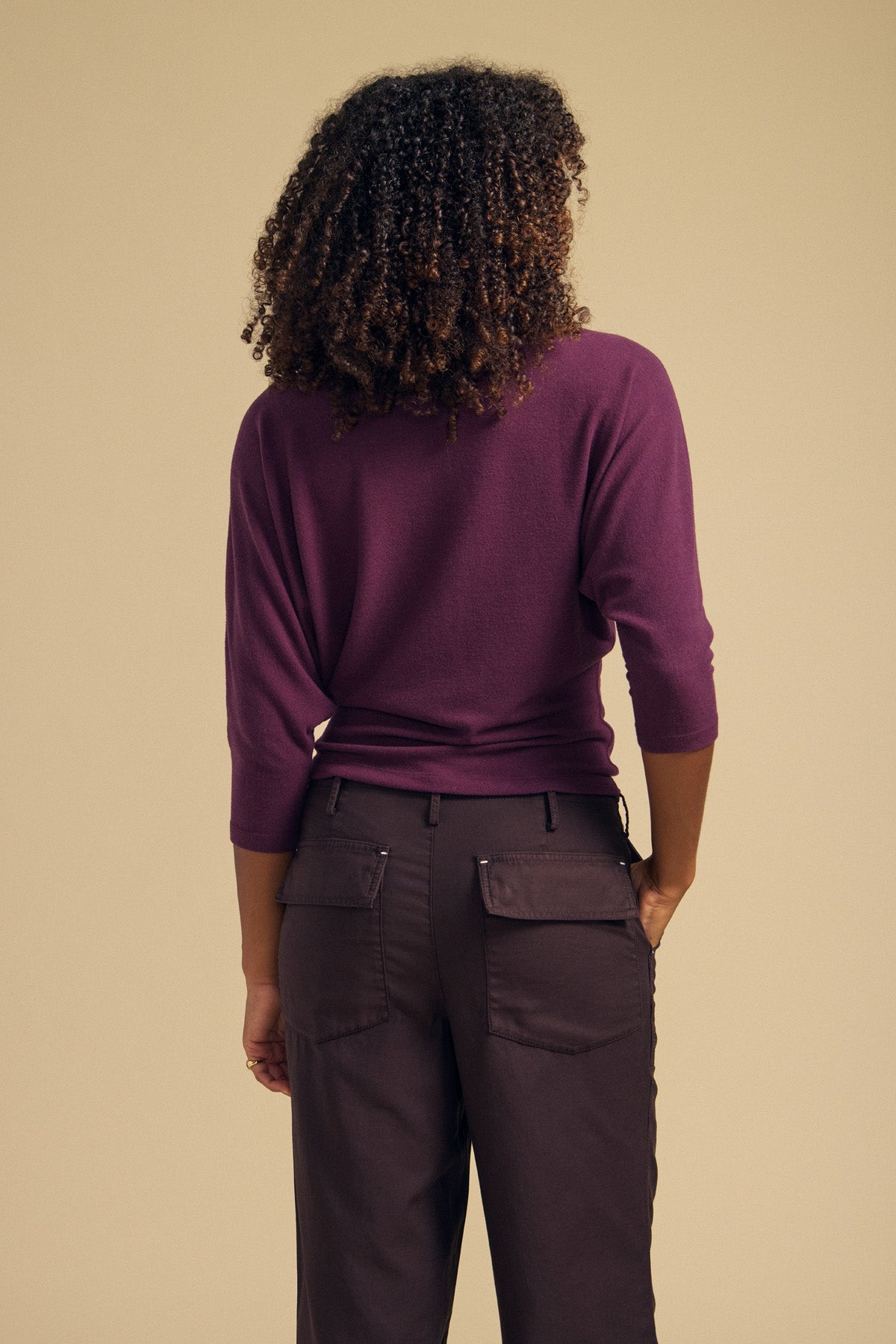 A person with curly hair is seen from behind, standing against a plain beige background, wearing the Velvet by Graham & Spencer CECILIA TOP in purple—a brushed knit long-sleeve top—paired with dark pants.-42063734669505