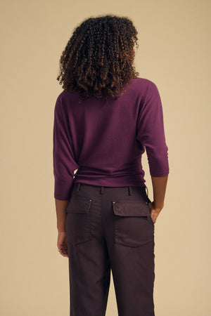 A person with curly hair is seen from behind, standing against a plain beige background, wearing the Velvet by Graham & Spencer CECILIA TOP in purple—a brushed knit long-sleeve top—paired with dark pants.