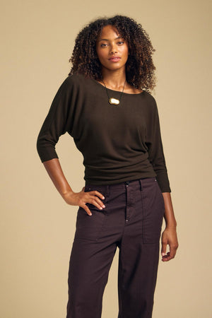 Woman wearing a dark brown, soft knit CECILIA TOP by Velvet by Graham & Spencer, featuring a boat-neck, three-quarter length dolman sleeves, and a gold pendant necklace.