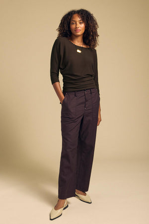 A person wearing a dark olive CECILIA TOP with a boat neck and dolman sleeves, dark trousers, and pointed-toe flats.