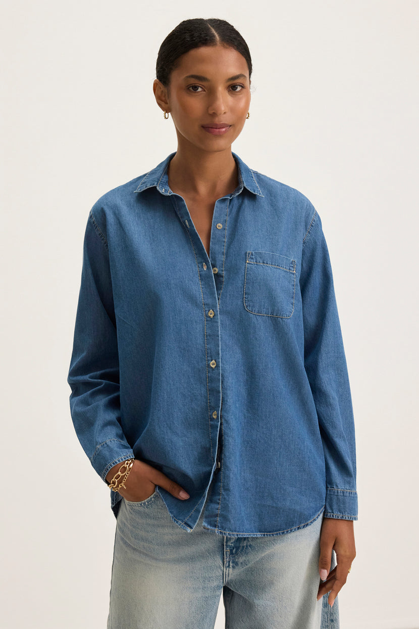 A person models the Velvet by Graham & Spencer KASSA CHAMBRAY SHIRT, featuring a button-up front, paired with light jeans against a plain background for a timeless look.