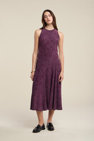A woman stands against a plain background, wearing the INGRID SILK DRESS by Velvet by Graham & Spencer—an ankle-length, sleeveless maroon dress with small floral patterns—paired with black loafers and a bracelet on her left wrist.