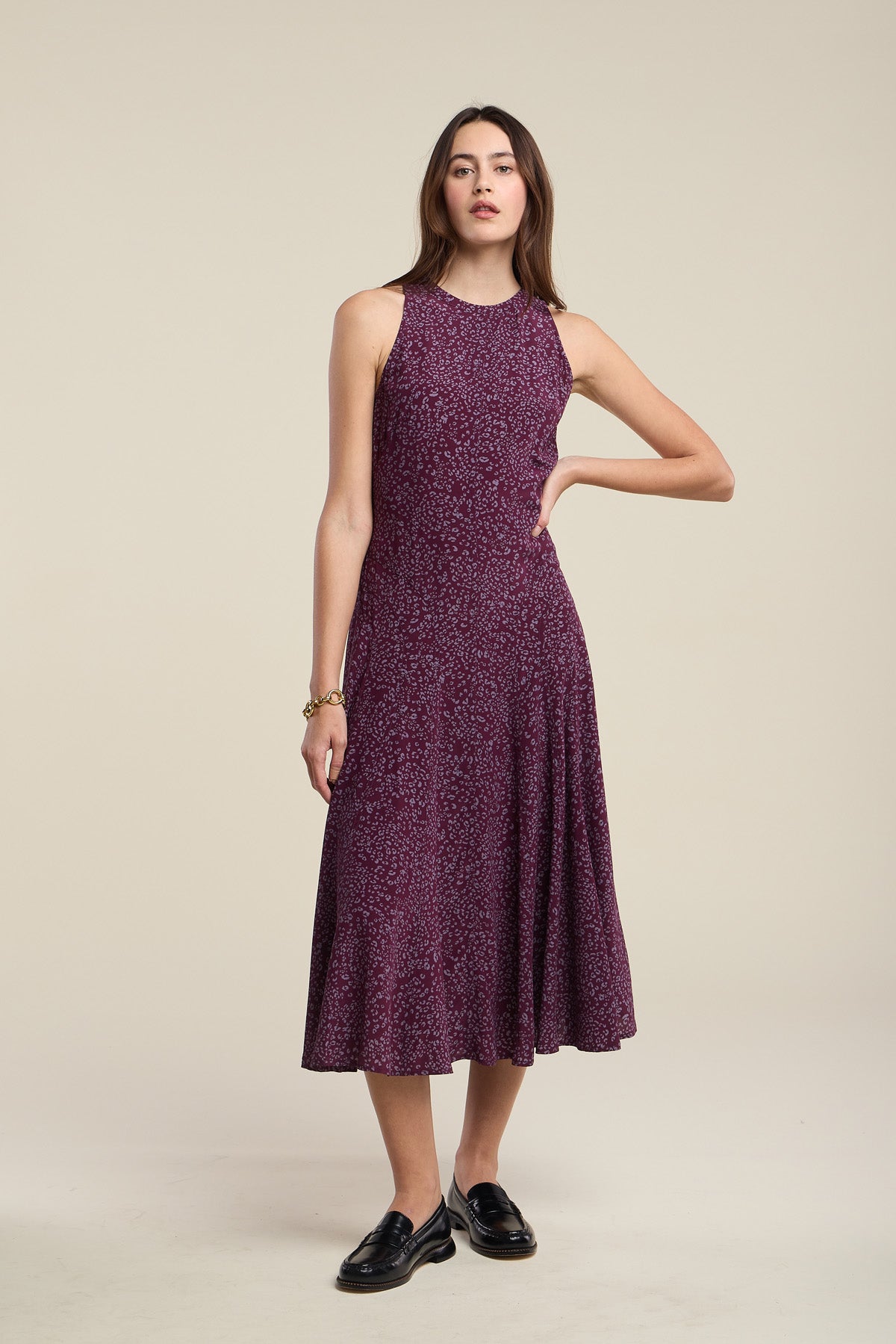 A woman stands against a neutral background wearing the Velvet by Graham & Spencer INGRID SILK DRESS—a sleeveless, knee-length purple patterned dress—paired with black loafers and one hand on her hip.
