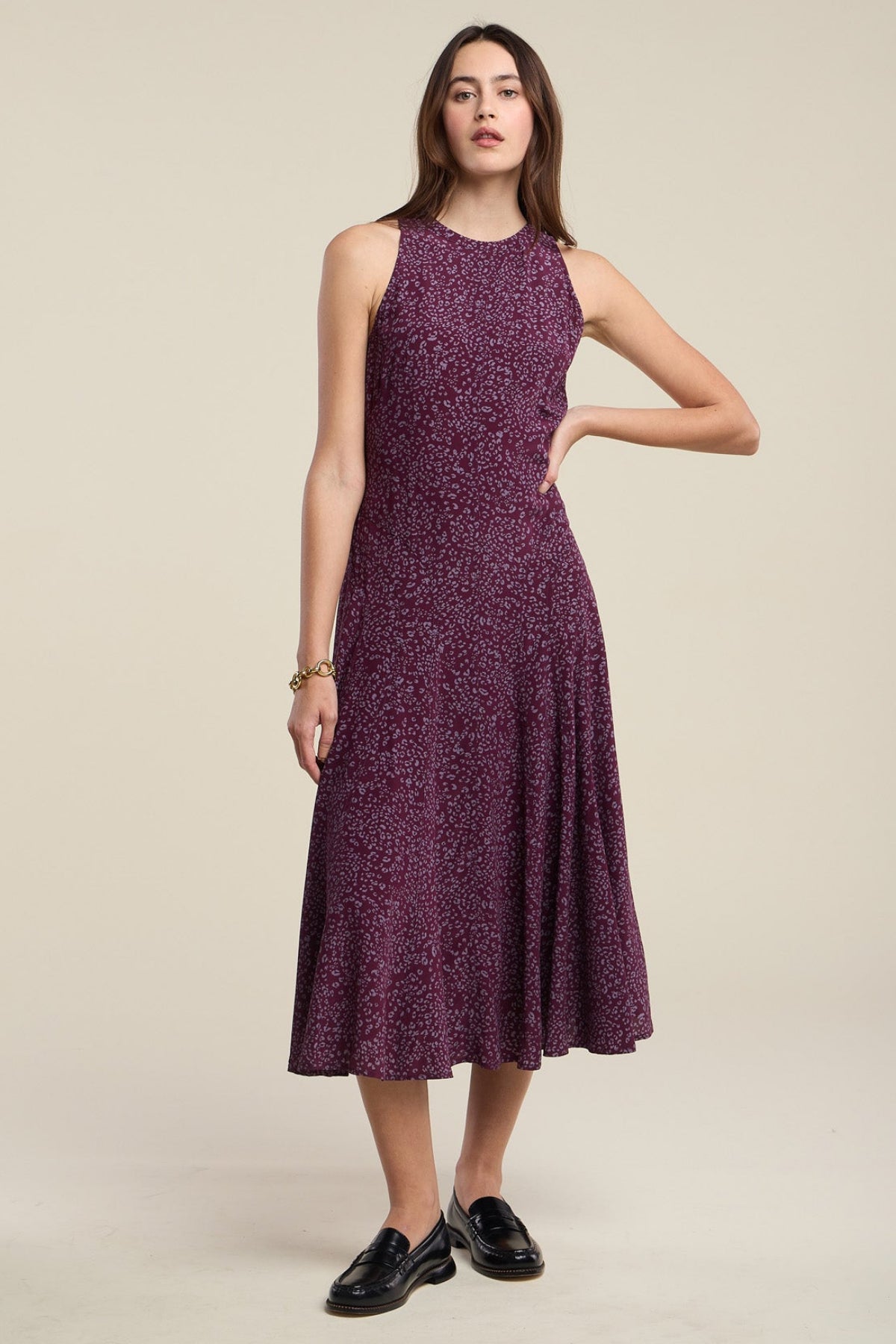 A woman stands against a neutral background wearing the Velvet by Graham & Spencer INGRID SILK DRESS—a sleeveless, knee-length purple patterned dress—paired with black loafers and one hand on her hip.-42063743484097