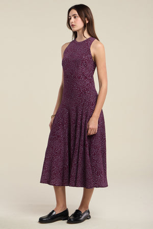 A woman stands sideways against a plain background, wearing the INGRID SILK DRESS—a sleeveless, purple patterned midi dress by Velvet by Graham & Spencer—paired with black loafers.