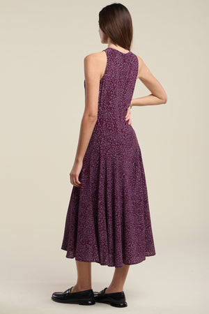 A woman stands with her back to the camera against a plain background, wearing the INGRID SILK DRESS by Velvet by Graham & Spencer—a sleeveless, ankle-length purple patterned dress—paired with black loafers.