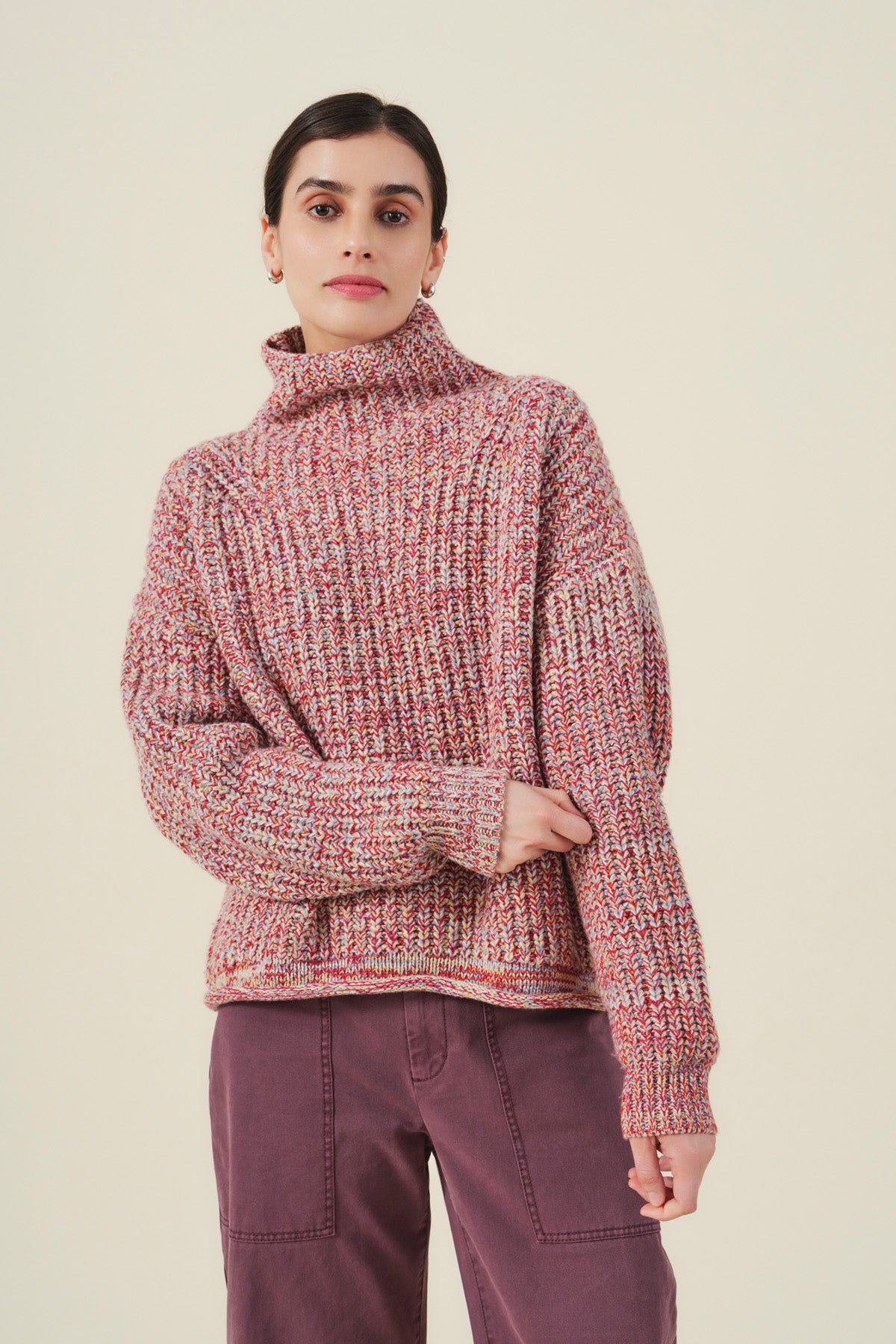A person wearing the DENNIE SWEATER by Velvet by Graham & Spencer—a marled knit chunky turtleneck in pink tones—paired with purple pants stands against a plain light background.-41970979274945