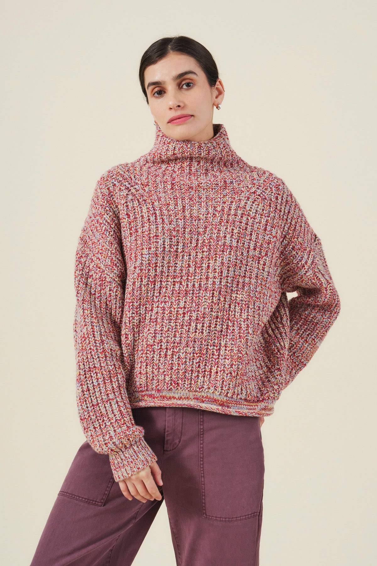 A person stands against a plain background wearing the DENNIE SWEATER, a chunky knit wool blend turtleneck by Velvet by Graham & Spencer, paired with purple pants and one hand resting on their hip.-41970979307713