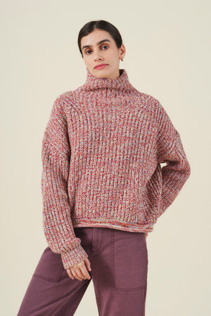 A person stands against a plain background wearing the DENNIE SWEATER, a chunky knit wool blend turtleneck by Velvet by Graham & Spencer, paired with purple pants and one hand resting on their hip.