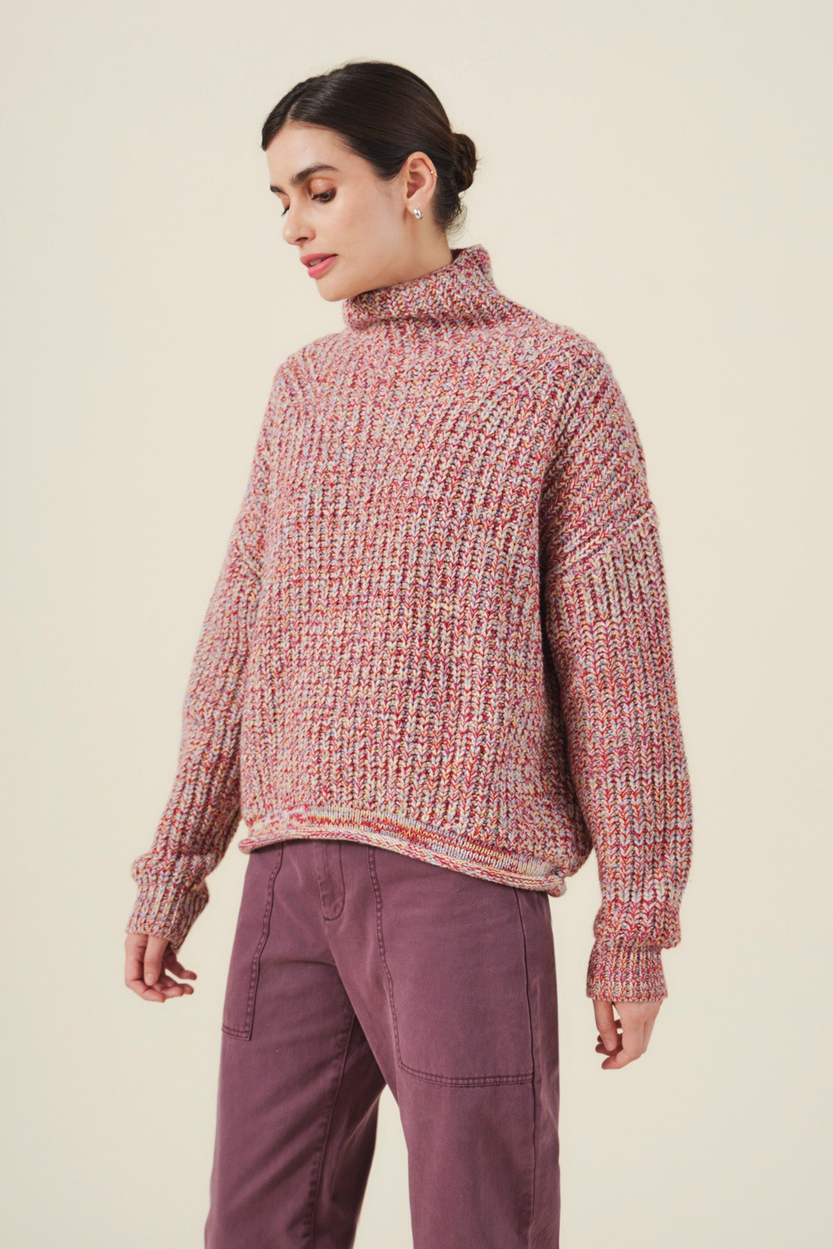 A woman stands against a plain background wearing the DENNIE SWEATER by Velvet by Graham & Spencer—a loose, chunky knit turtleneck in pink tones—paired with mauve pants. The cozy wool-blend sweater brings textured warmth to her effortlessly stylish look.