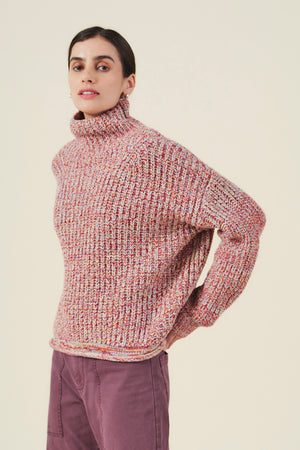 A person wearing the DENNIE SWEATER by Velvet by Graham & Spencer—a chunky knit turtleneck in multicolored hues—paired with mauve pants, stands with hands on hips against a plain light background.