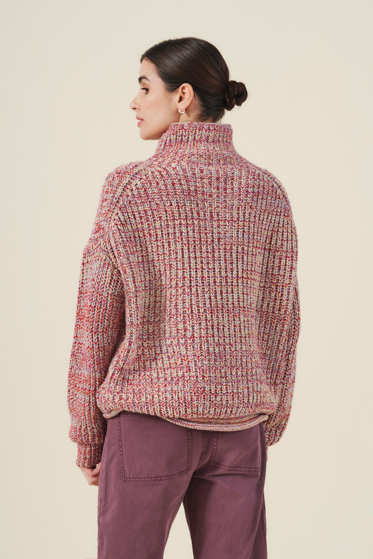 A woman stands facing away, wearing the DENNIE SWEATER by Velvet by Graham & Spencer—a cozy, chunky knit in multicolored hues with a turtleneck—paired with lilac pants, her dark hair pulled back in a low bun.