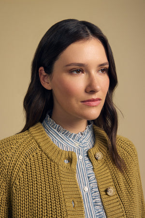 A woman with long brown hair models the Velvet by Graham & Spencer LISABETH CARDIGAN, a chunky cropped style layered over a blue striped shirt, standing against a plain beige background.