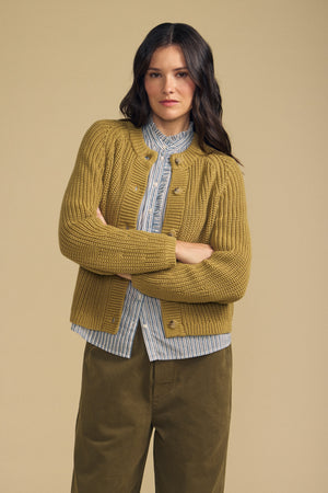 A woman stands with arms crossed, wearing the Velvet by Graham & Spencer LISABETH CARDIGAN over a striped shirt and olive-green pants, against a plain beige background.