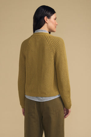 A person stands with their back to the camera, wearing the LISABETH CARDIGAN by Velvet by Graham & Spencer—a chunky cropped layer styled over a striped shirt and olive pants—set against a plain beige background for an effortlessly stylish transitional look suitable for any season.