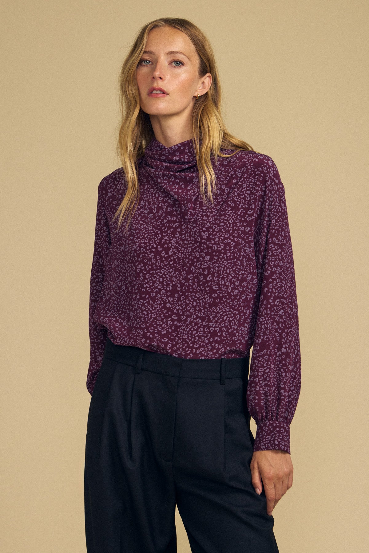 A woman wearing the NALA SILK BLOUSE by Velvet by Graham & Spencer—featuring a purple cheetah print, long sleeves, and a high neckline—paired with black trousers, stands against a beige background.