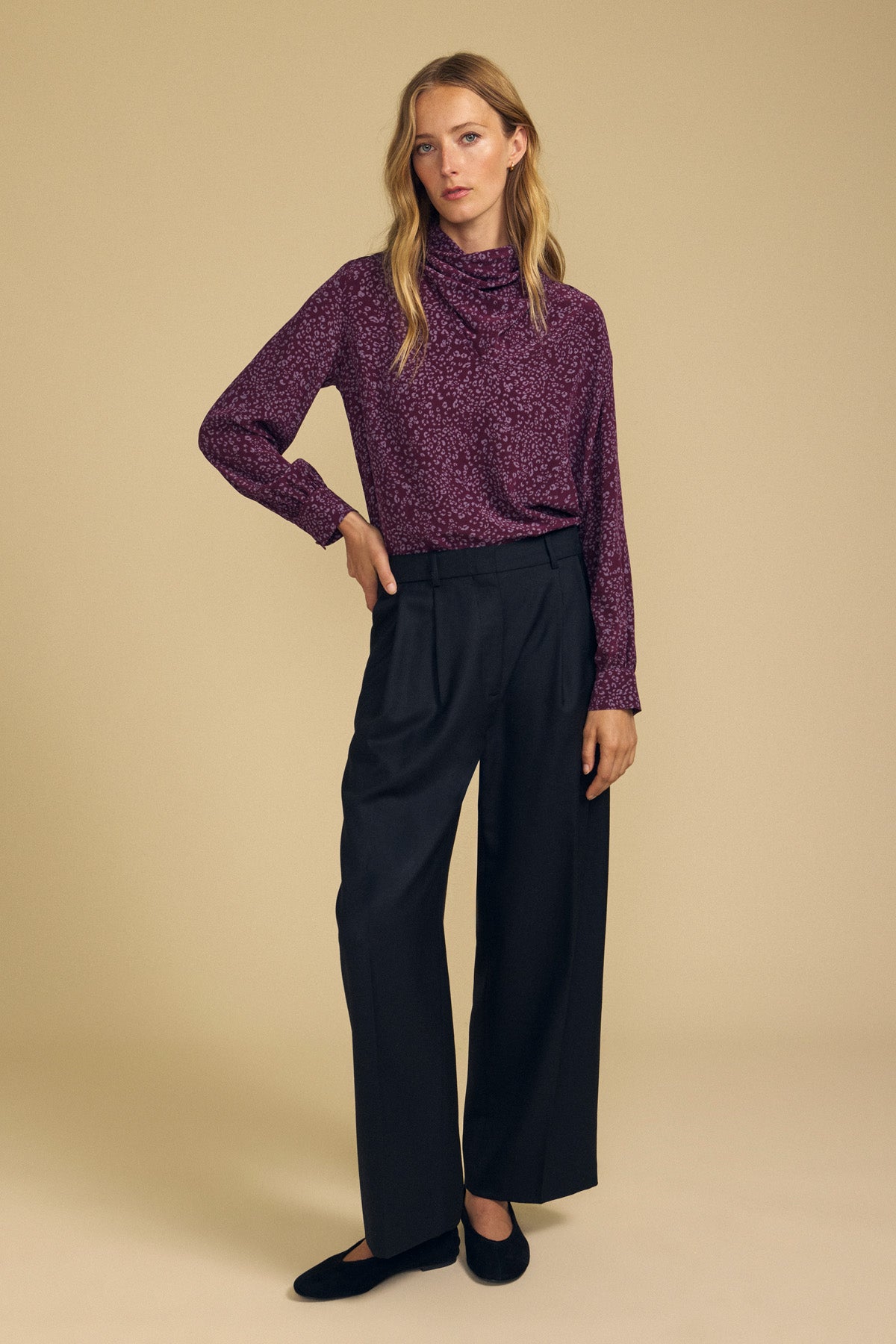 A person stands against a beige background wearing the NALA SILK BLOUSE by Velvet by Graham & Spencer—a long-sleeved, purple cheetah-printed blouse with a cowl neckline—paired with high-waisted black wide-leg pants and black shoes.-42063824519361