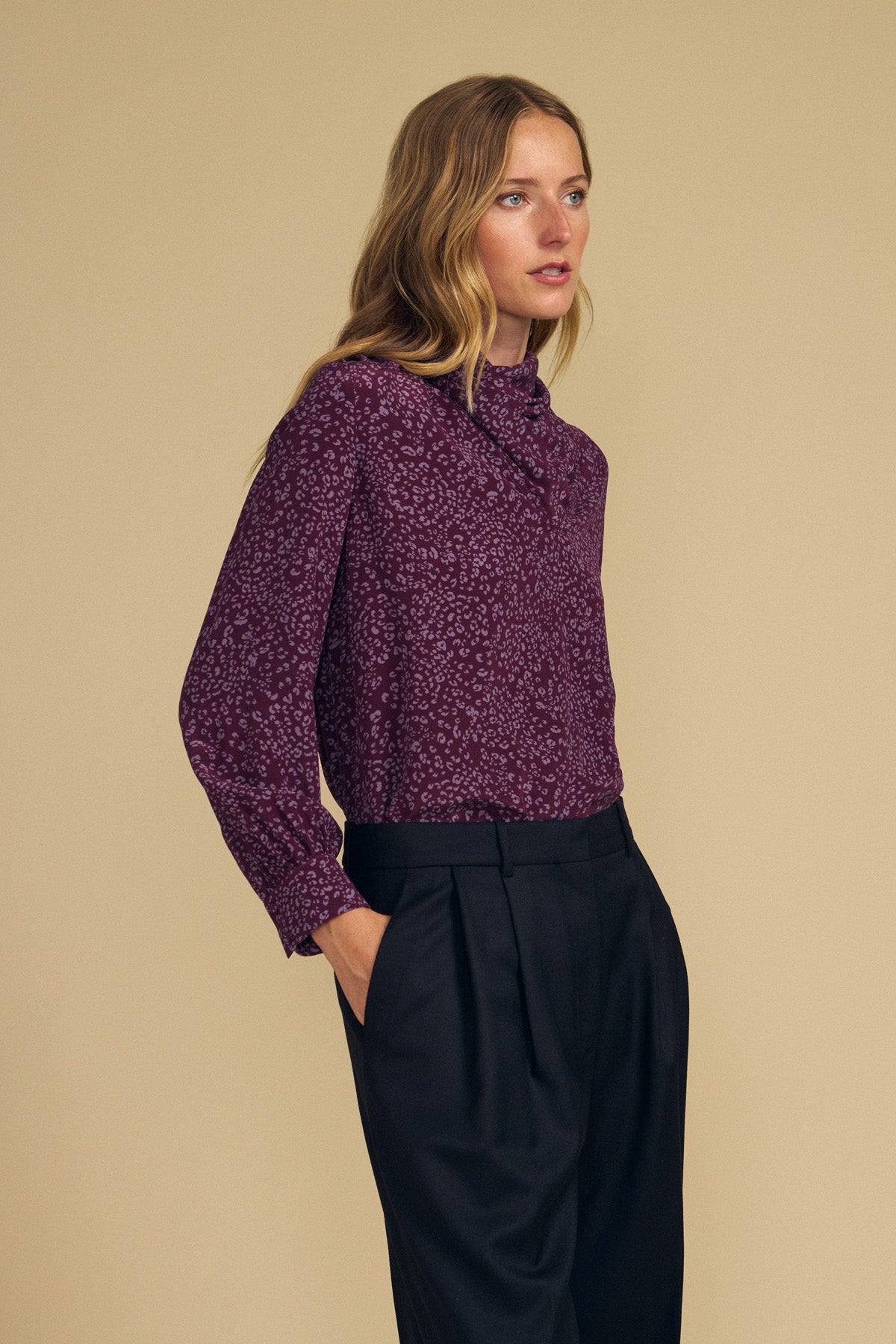 A woman stands against a beige background, wearing the NALA SILK BLOUSE by Velvet by Graham & Spencer—a purple cheetah-printed, cowl-neck blouse—paired with black pleated trousers and her hands in her pockets.