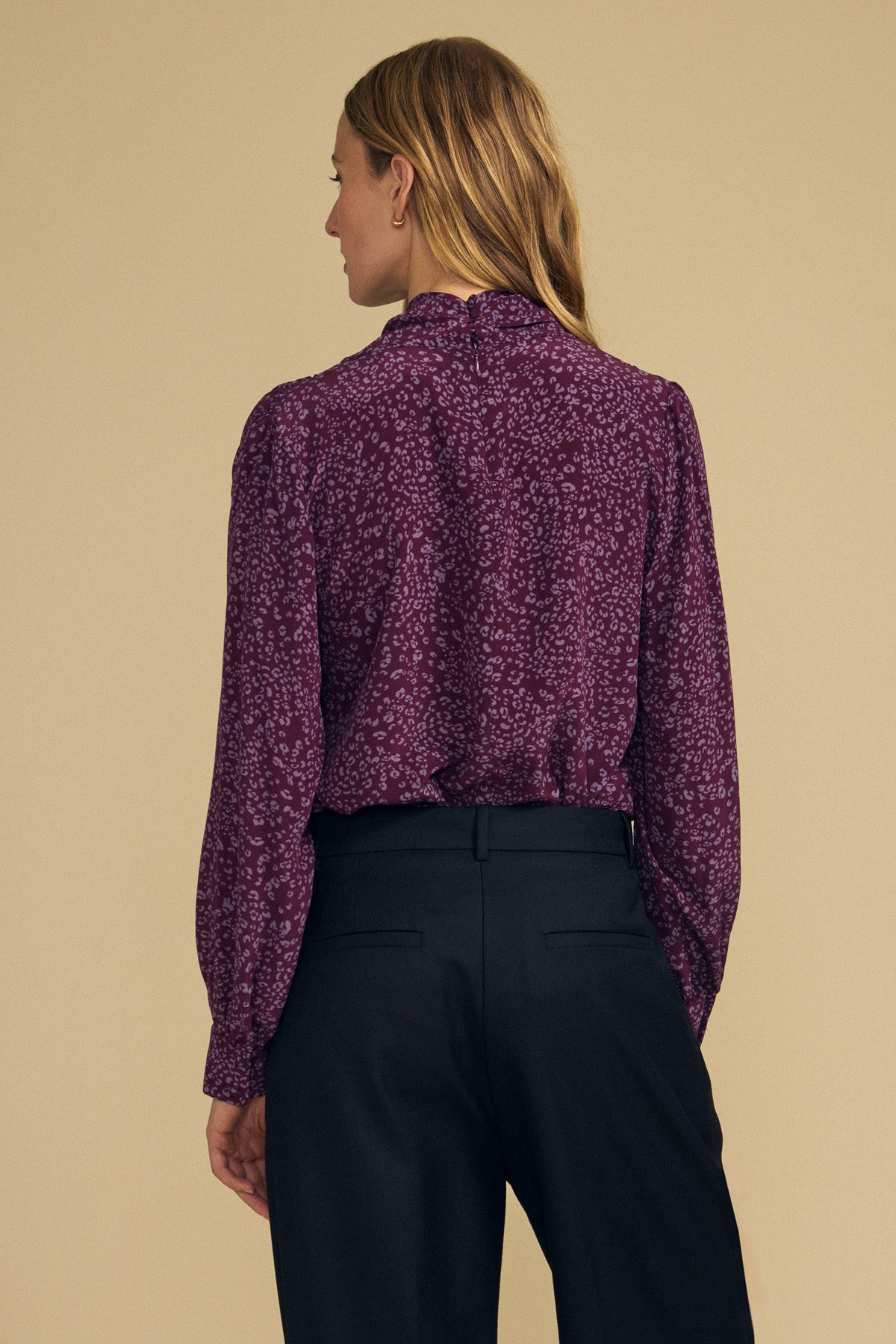 A woman stands with her back to the camera, modeling the NALA SILK BLOUSE by Velvet by Graham & Spencer—a long-sleeve, cheetah print blouse featuring a cowl neckline—paired with black pants against a plain beige background.