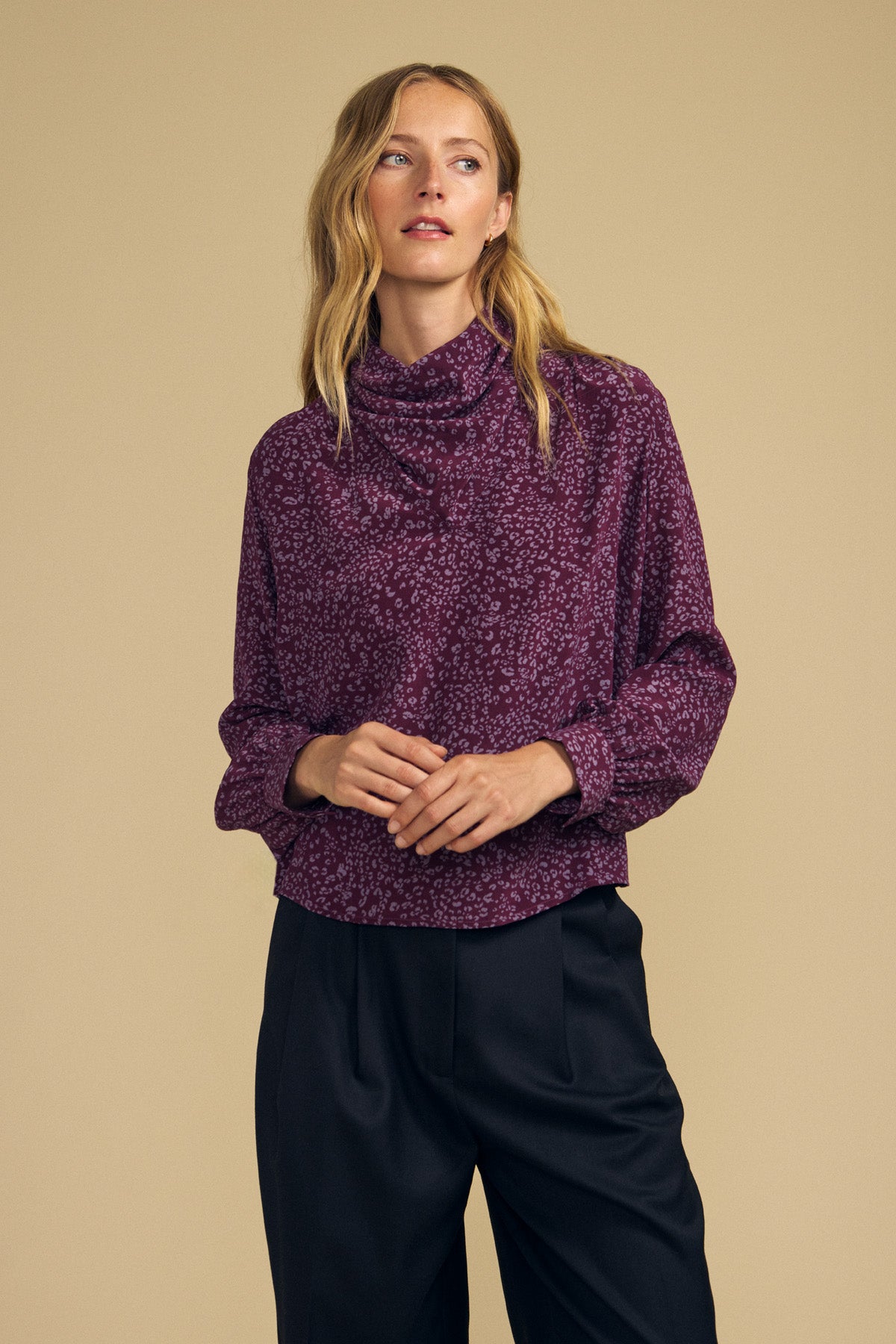 A woman wearing the NALA SILK BLOUSE by Velvet by Graham & Spencer—a long-sleeved, cheetah-printed top with a high collar—paired with black pants, stands against a plain beige background.