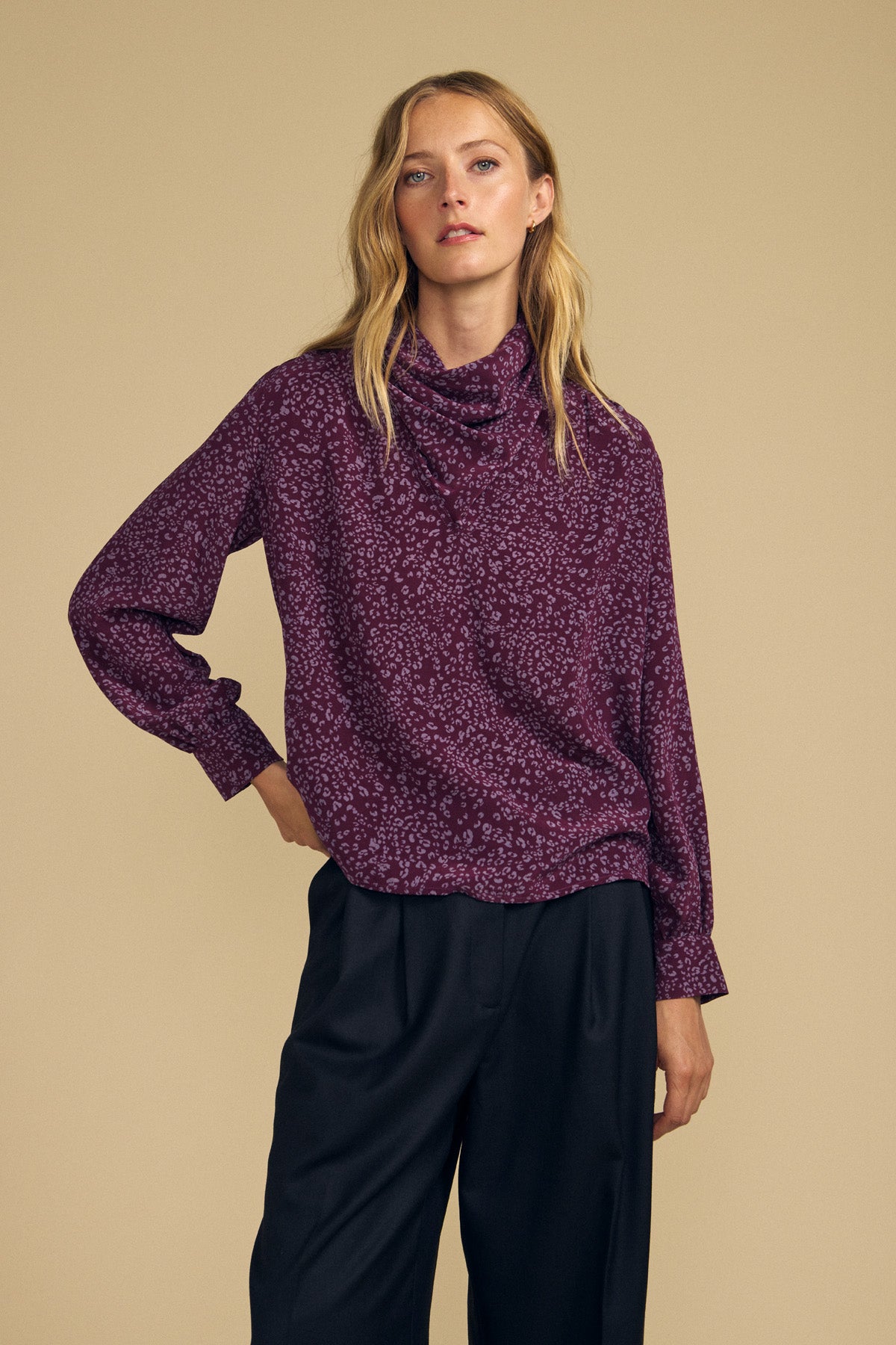 A person with long blonde hair wears the NALA SILK BLOUSE by Velvet by Graham & Spencer—a purple, cheetah-printed blouse with long sleeves—paired with loose black trousers, standing against a beige background.