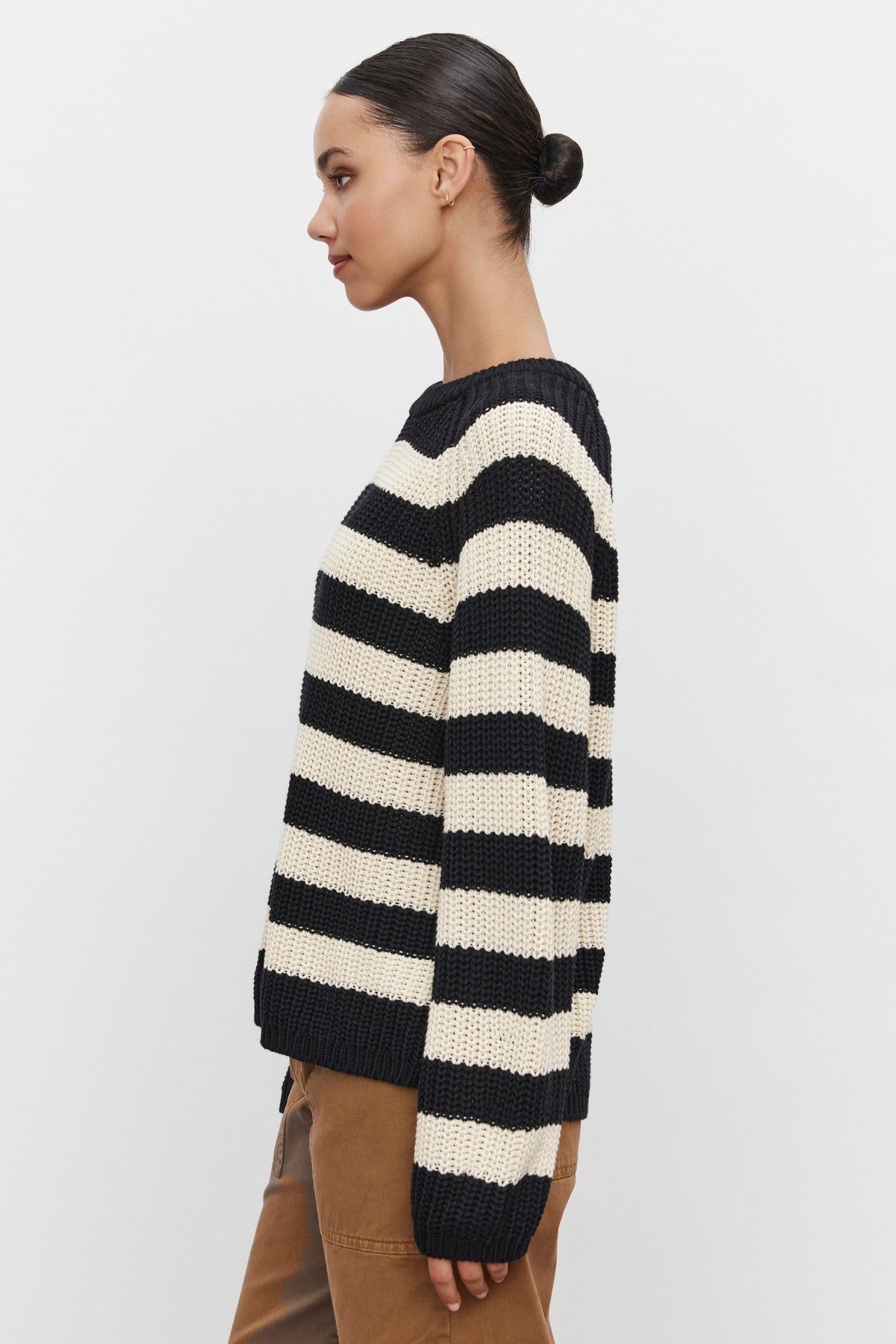   A woman stands in profile wearing the OLLIE SWEATER by Velvet by Graham & Spencer, a black and white striped pattern sweater with a crew neckline, paired with brown pants. Her hair is tied back in a bun. 