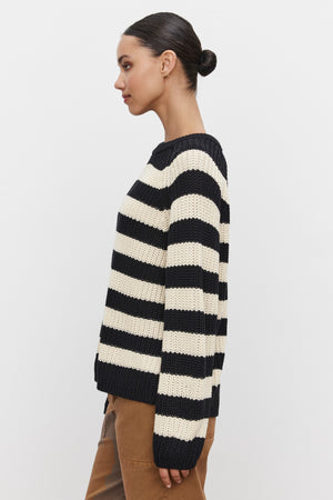 A woman stands in profile wearing the OLLIE SWEATER by Velvet by Graham & Spencer, a black and white striped pattern sweater with a crew neckline, paired with brown pants. Her hair is tied back in a bun.