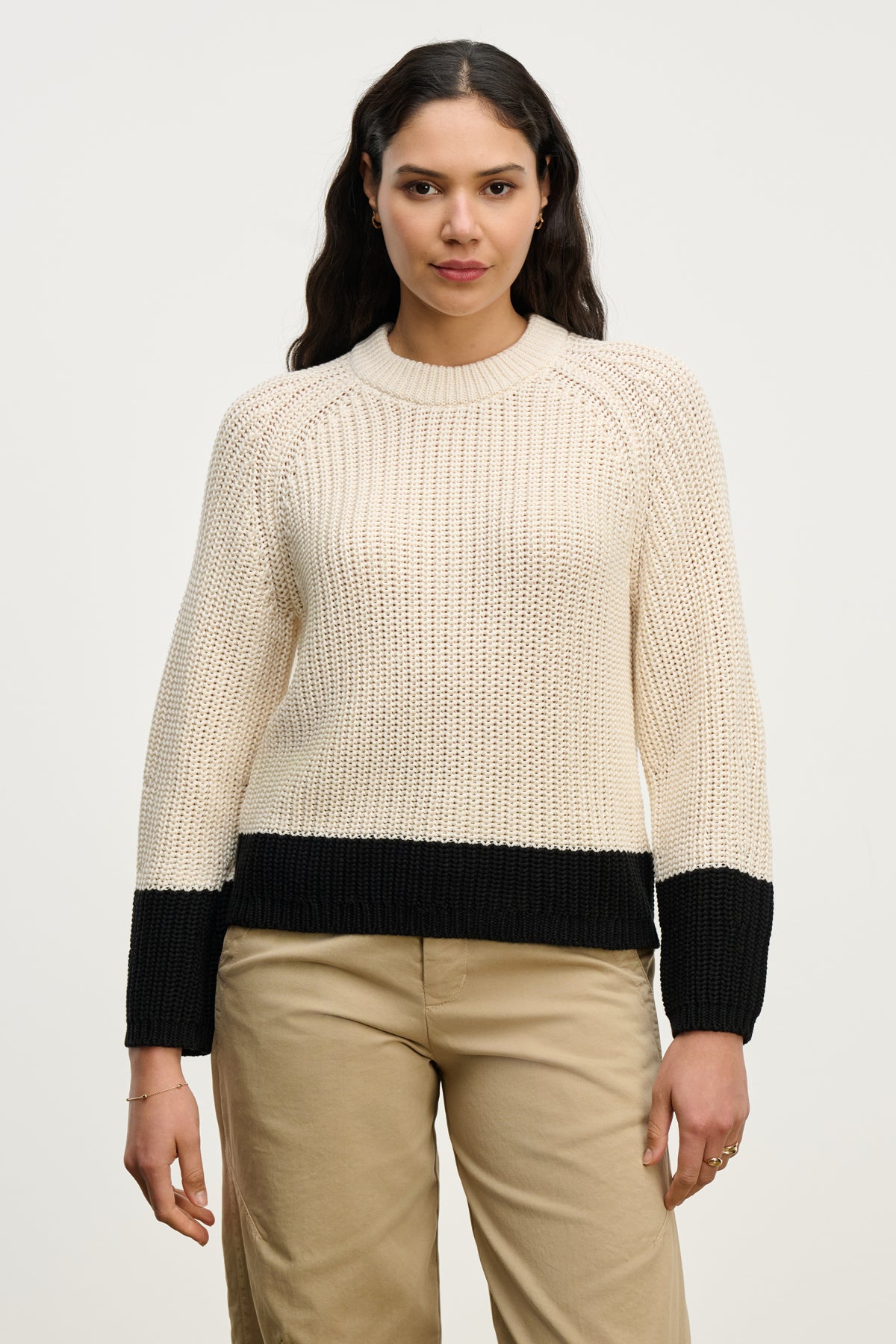 A woman faces forward in the SHAY SWEATER by Velvet by Graham & Spencer—a beige color block knit with black cuffs, hem, and crew neckline—paired with beige pants against a plain white background.