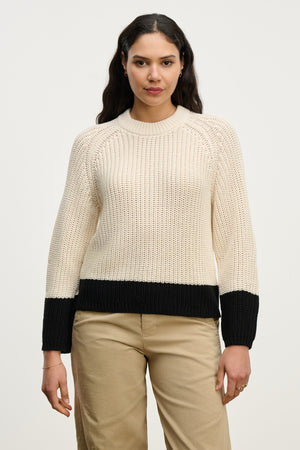 A woman faces forward in the SHAY SWEATER by Velvet by Graham & Spencer—a beige color block knit with black cuffs, hem, and crew neckline—paired with beige pants against a plain white background.
