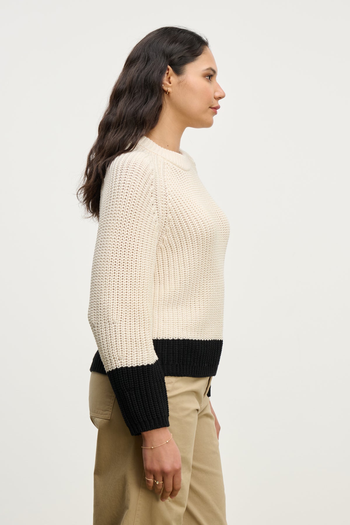 A woman stands in profile against a plain background wearing the SHAY SWEATER by Velvet by Graham & Spencer, featuring a cream color block design with black cuffs and hem, crew neckline, and paired with beige pants.