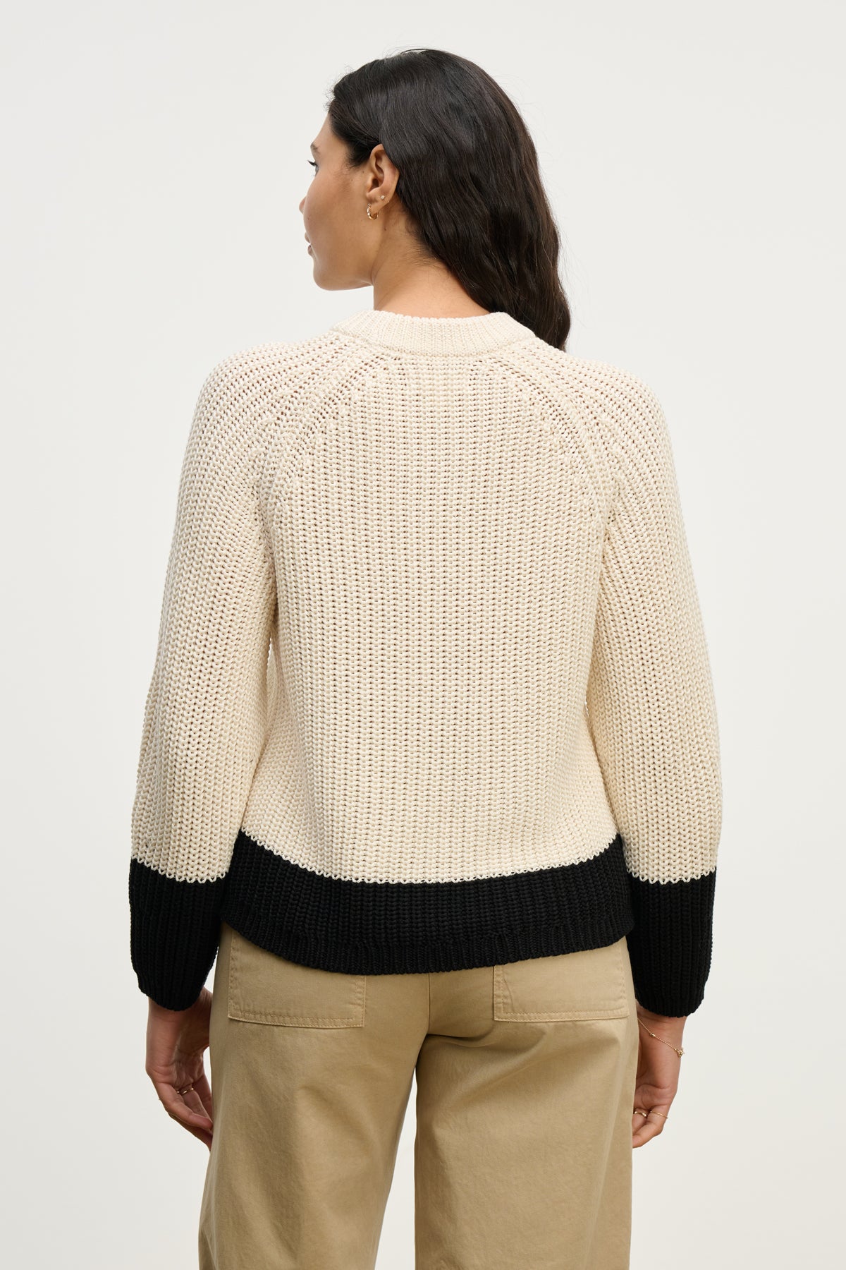 A woman stands facing away, wearing the Velvet by Graham & Spencer SHAY SWEATER—a cozy color block knit with black cuffs and hem—paired with beige pants against a plain background.-40046145929409