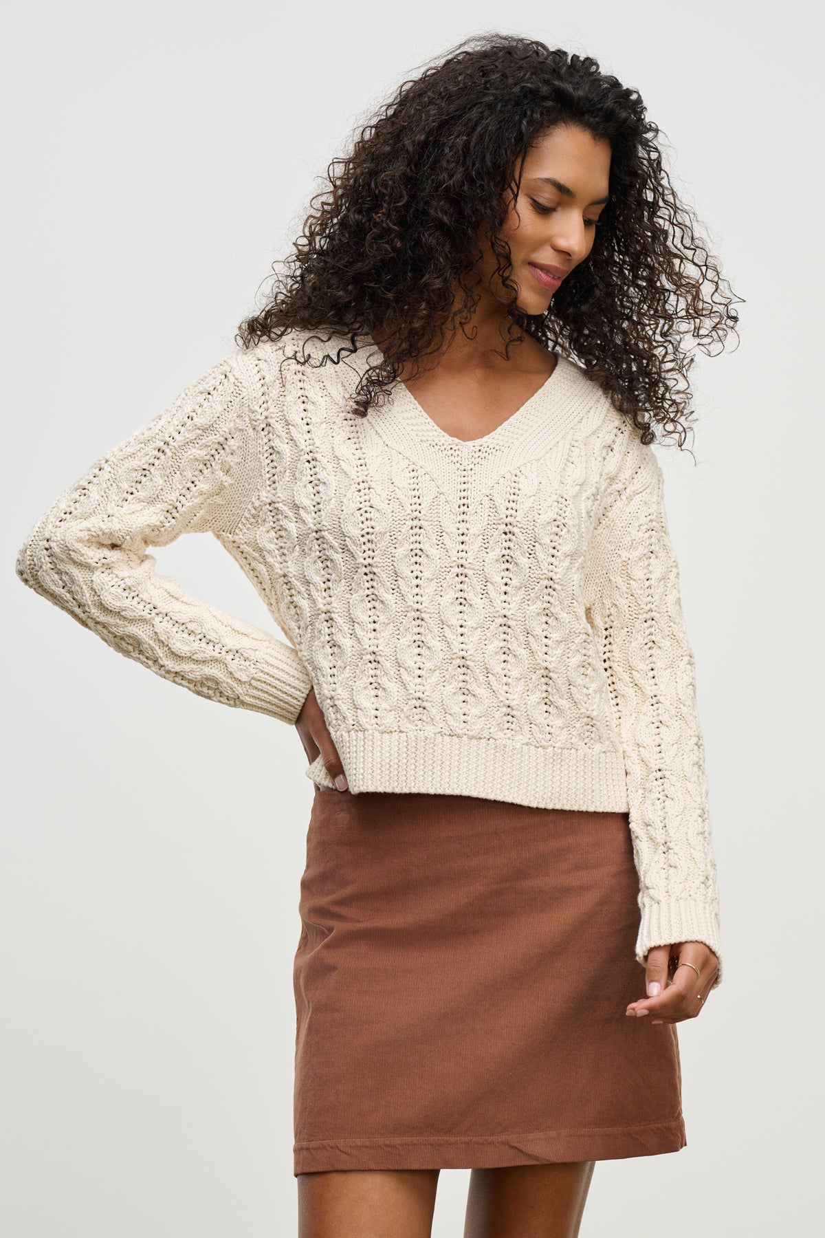 A woman with curly hair wears the JOYCE SWEATER by Velvet by Graham & Spencer, a chunky Pima cotton cable knit, paired with a brown skirt against a plain light background.-39800333992129