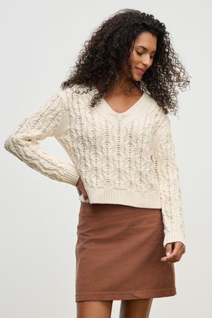 A woman with curly hair wears the JOYCE SWEATER by Velvet by Graham & Spencer, a chunky Pima cotton cable knit, paired with a brown skirt against a plain light background.