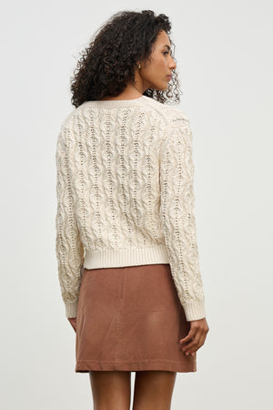 A woman stands sideways against a plain light background, wearing the JOYCE SWEATER by Velvet by Graham & Spencer—a cream-colored, boxy fit chunky Pima cotton sweater—paired with a brown suede skirt.