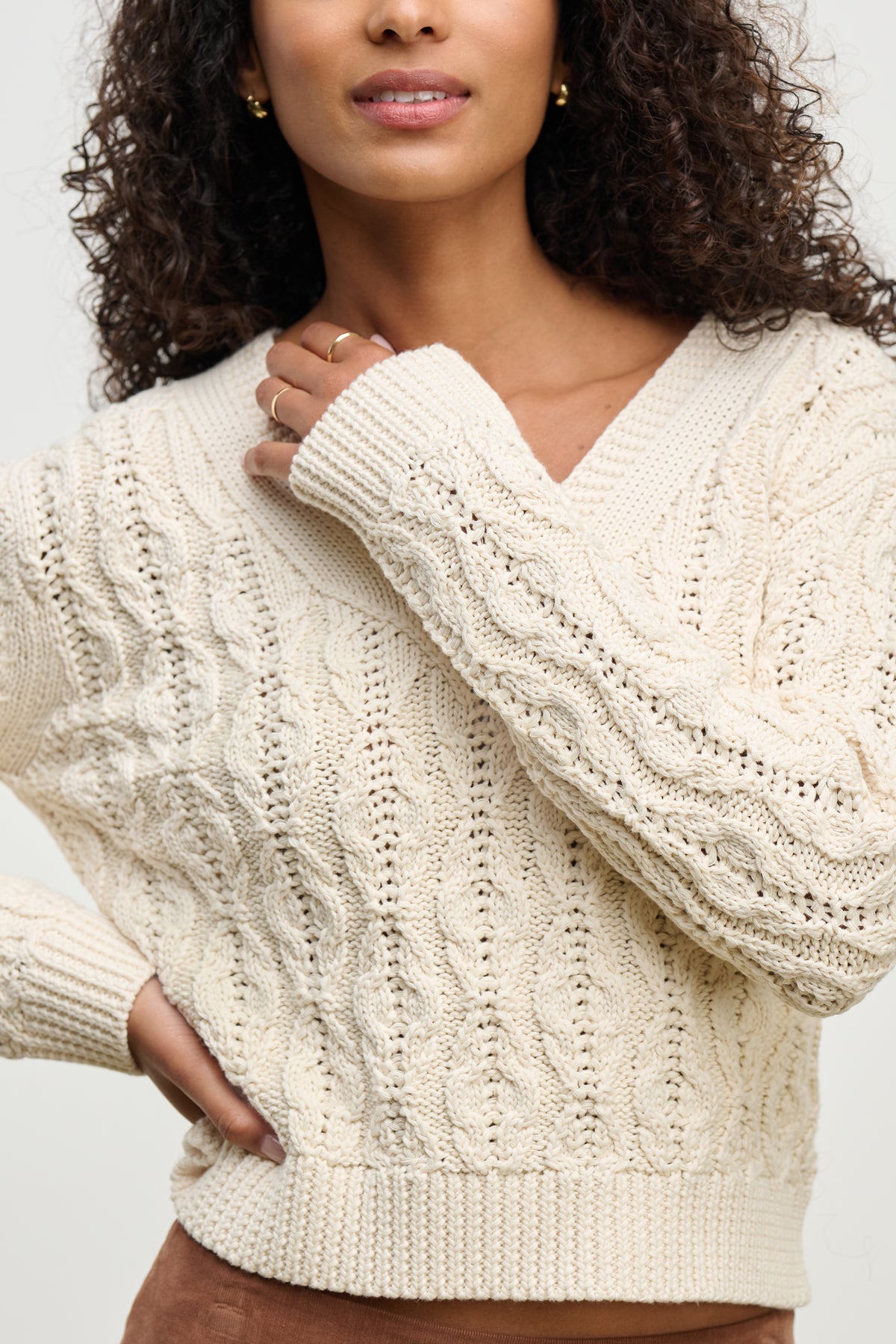   A person wears the JOYCE SWEATER by Velvet by Graham & Spencer—a cream chunky Pima cotton cable knit—with one hand near their chin and the other on their hip. 