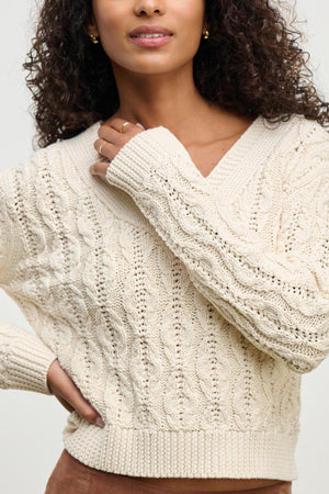 A person wears the JOYCE SWEATER by Velvet by Graham & Spencer—a cream chunky Pima cotton cable knit—with one hand near their chin and the other on their hip.
