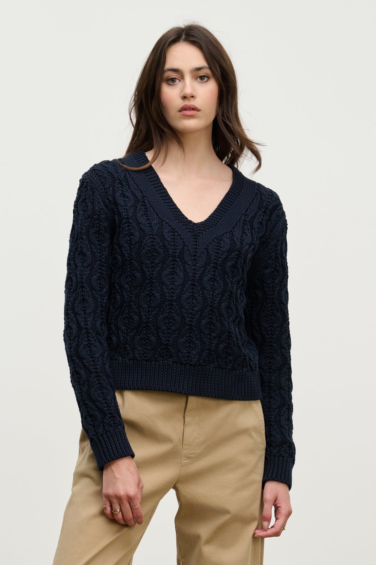 A woman wears the JOYCE SWEATER by Velvet by Graham & Spencer, a navy chunky Pima cotton cable knit with a boxy fit, paired with beige pants against a plain light background.-39814892912833