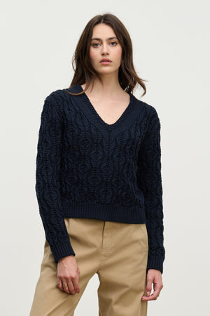 A woman wears the JOYCE SWEATER by Velvet by Graham & Spencer, a navy chunky Pima cotton cable knit with a boxy fit, paired with beige pants against a plain light background.