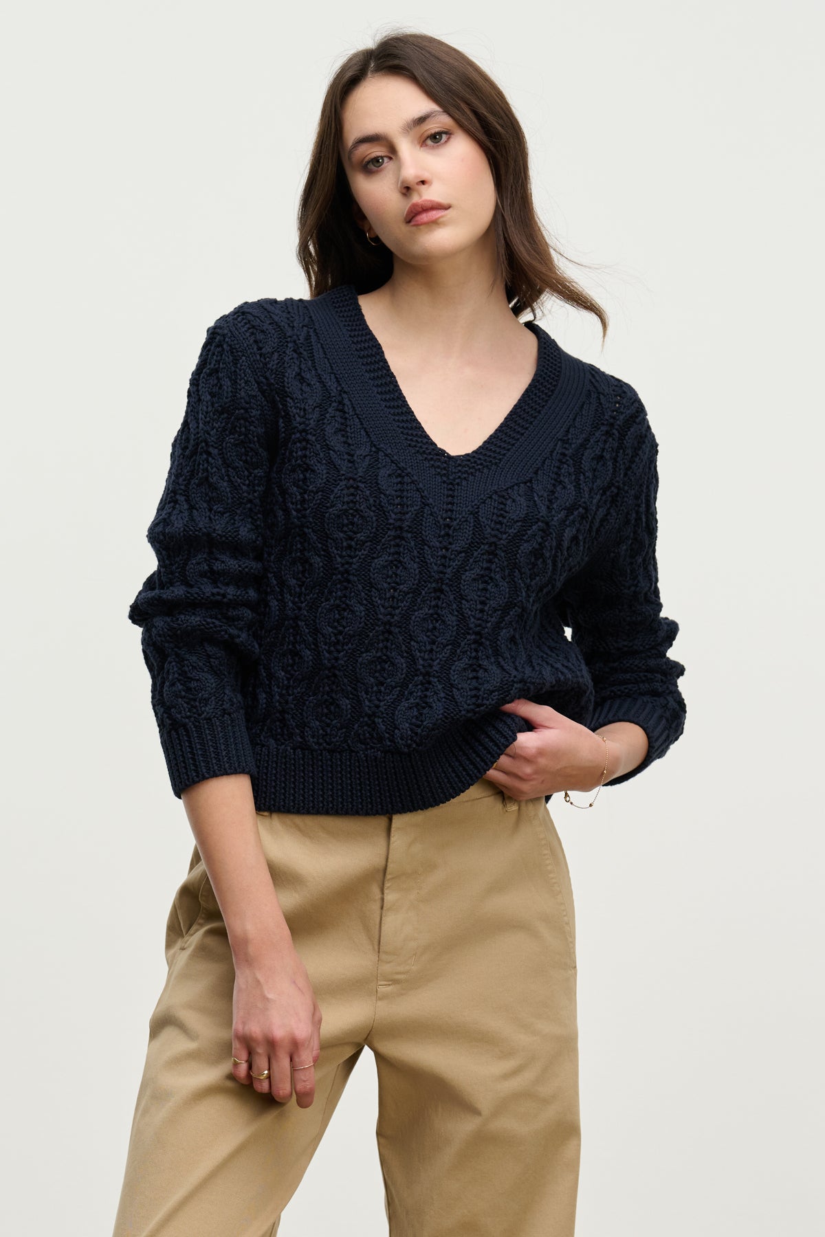 A woman wears the JOYCE SWEATER by Velvet by Graham & Spencer—a dark blue, chunky Pima cotton cable knit with a boxy fit—paired with beige pants, standing against a plain light background.-39814892880065