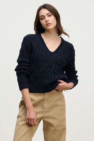 A woman wears the JOYCE SWEATER by Velvet by Graham & Spencer—a dark blue, chunky Pima cotton cable knit with a boxy fit—paired with beige pants, standing against a plain light background.