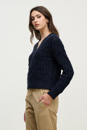 A woman stands with one hand in her pocket, wearing the Velvet by Graham & Spencer JOYCE SWEATER— a navy boxy fit cable knit made from chunky Pima cotton— and beige pants, against a plain light background.