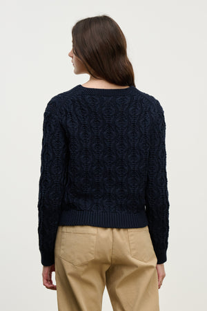 A woman with long brown hair is seen from the back wearing the JOYCE SWEATER by Velvet by Graham & Spencer in navy chunky Pima cotton cable knit, paired with beige pants, against a plain light background.